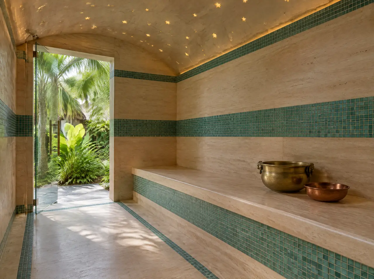 Interior of a small hammam in a Koh Samui spa hotel: pale travertine bench and walls with bands of lagoon-teal mosaic, a sloped vaulted ceiling with points of light and a glass door open to a palm garden