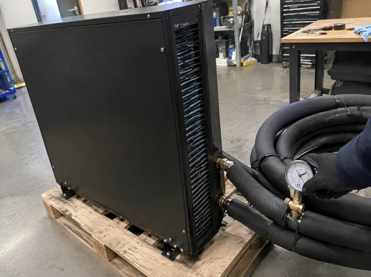 Plain black rectangular chiller casing on a pallet in a workshop with black insulated pipes and a technician's gloved hand on a pressure gauge