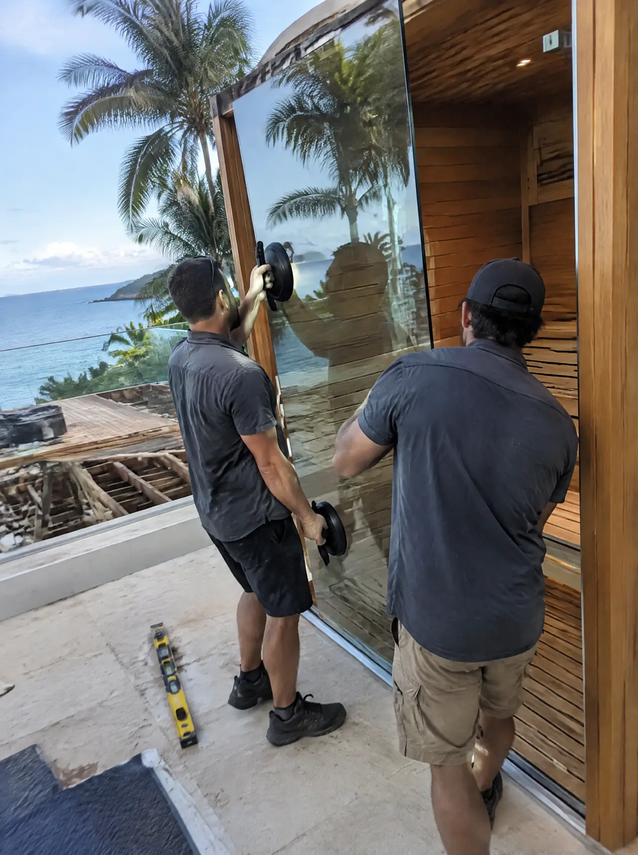 Two installers lifting a tempered glass panel with suction cups into a teak sauna frame on a villa terrace